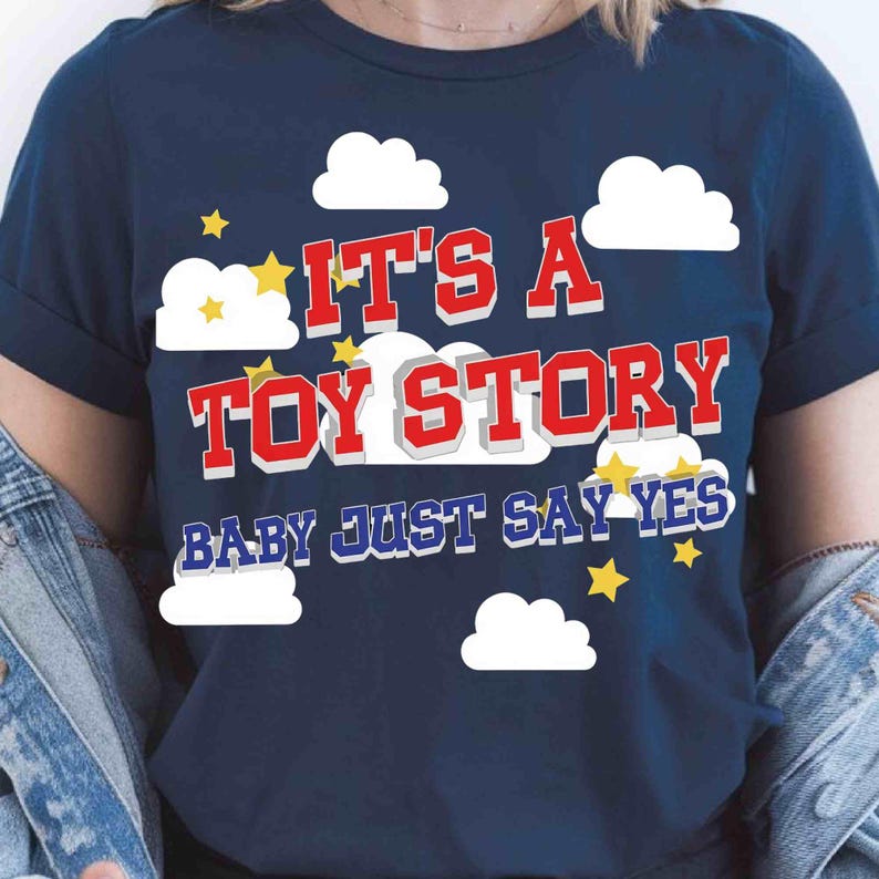 Puede incluir: Una camiseta azul marino con un estampado gr&aacute;fico de nubes blancas y estrellas amarillas. El texto IT'S A TOY STORY aparece en letras rojas con borde blanco, seguido del texto azul BABY JUST SAY YES en una fuente negrita sin remates.