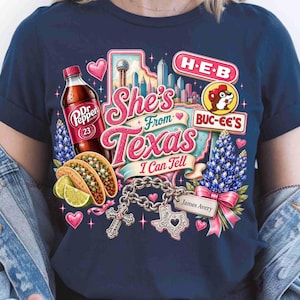 May include: A navy blue t-shirt featuring a graphic print with the text She's From Texas I Can Tell. The design includes a Dr Pepper bottle, tacos with lime wedges, a city skyline, H-E-B and Buc-ee's logos, bluebonnets, and a charm bracelet with a cross.