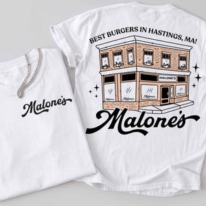 May include: Two white t-shirts. One shirt has the word "Malone's" in a cursive font. The other shirt has a graphic of a two-story building with the text "BEST BURGERS IN HASTINGS, MA!" and "Malone's" in a cursive font.