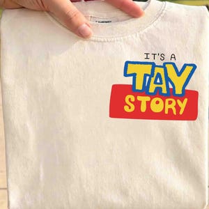 May include: A cream-colored t-shirt featuring a graphic print on the chest. The text reads IT'S A TAY STORY, with TAY in large blue letters with yellow outlines and STORY in yellow letters on a red rectangular background. The shirt has a crew neckline.