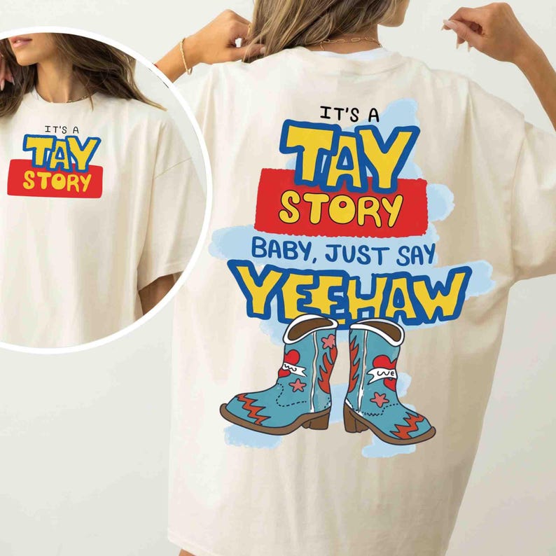 May include: An off-white t-shirt featuring a graphic print on the back and a smaller version on the front. The design includes the text IT'S A TAY STORY BABY, JUST SAY YEEHAW in blue, yellow, and red, accompanied by two light blue cowboy boots.