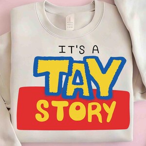 May include: A cream-colored crewneck sweatshirt featuring the text IT'S A TAY STORY. The word TAY is printed in large yellow letters with a thick blue outline, while the word STORY is printed in yellow on a solid red rectangular background.