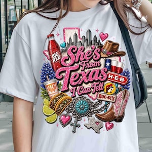 May include: A white t-shirt featuring a graphic print with the text She's From Texas I Can Tell. The design includes a Texas map, a city skyline, a Big Red bottle, a cowboy hat, a Texas flag, H-E-B logo, tacos, lime slices, a turquoise concho, a cowboy boot, and a Buc-ee's logo.