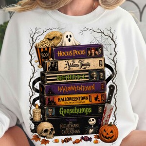 Könnte beinhalten: Ein weißes Sweatshirt mit einem Grafikdruck eines Stapels von VHS-Kassetten mit den Titeln Hocus Pocus, The Addams Family, Beetlejuice, Halloweentown, Halloweentown II, Goosebumps und The Nightmare Before Christmas, dazu ein Geist, ein Totenkopf und ein Kürbis.