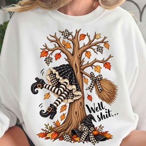 May include: A white sweatshirt featuring a graphic of a tree with a witch's legs and broomstick stuck in the branches. The design includes black and white checkered patterns, orange autumn leaves, a black witch hat, and the text Well shit...