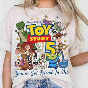 May include: A light-colored t-shirt featuring a colorful Toy Story 5 graphic with characters including Rex, Buzz Lightyear, Woody, Jessie, Bullseye, Forky, and others. The text reads Toy Story 5 and You're Got Friend In Me in blue script.