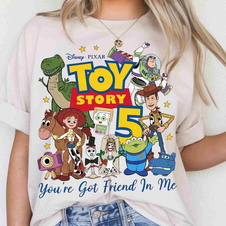 May include: A light-colored t-shirt featuring a colorful Toy Story 5 graphic with characters including Rex, Buzz Lightyear, Woody, Jessie, Bullseye, Forky, and others. The text reads Toy Story 5 and You're Got Friend In Me in blue script.