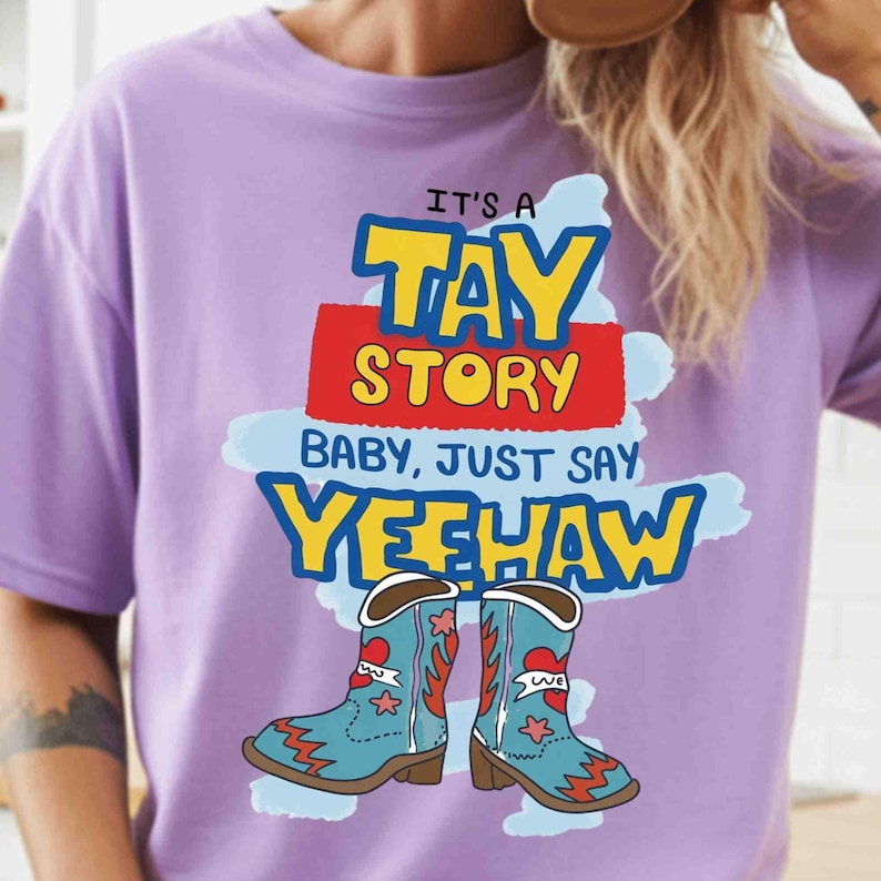 May include: A light purple t-shirt featuring a graphic print with the text IT'S A TAY STORY BABY, JUST SAY YEEHAW in yellow, red, and blue. Below the text are two light blue cowboy boots with red heart and star accents on a light blue background.