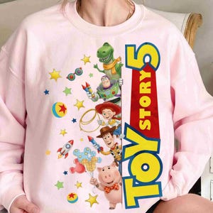 May include: A light pink crewneck sweatshirt featuring a graphic print of Toy Story 5 characters including Rex, Buzz Lightyear, Jessie, Woody, and Hamm. The design includes colorful stars, rockets, a ball, and the text Toy Story 5 in large letters.