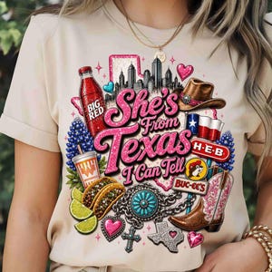 May include: A beige t-shirt featuring a graphic print with the text "She's From Texas I Can Tell". The design includes a skyline, a Big Red bottle, a cowboy hat, a Texas flag, H-E-B and Buc-ee's logos, tacos, lime slices, a turquoise concho, and a boot.