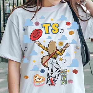 May include: A white t-shirt featuring a graphic of a person with long hair wearing a yellow plaid shirt and cow-print trousers, holding a guitar. The design includes a red hat, blue clouds, flowers, musical notes, yellow stars, and blocks with the text TS.