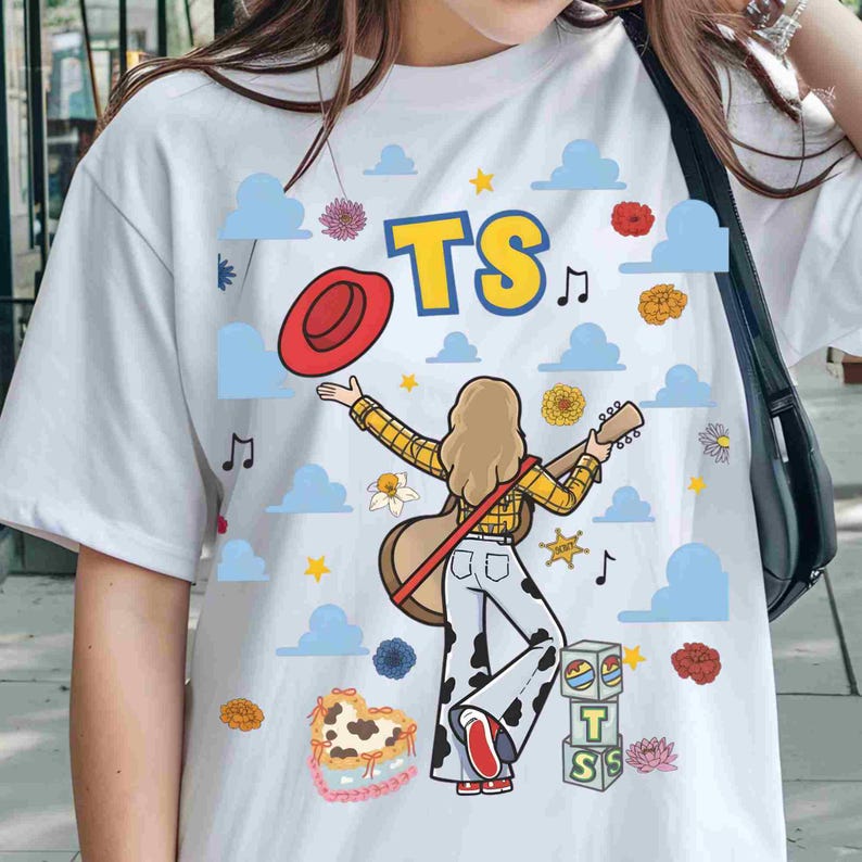 May include: A white t-shirt featuring a graphic of a person with long hair wearing a yellow plaid shirt and cow-print trousers, holding a guitar. The design includes a red hat, blue clouds, flowers, musical notes, yellow stars, and blocks with the text TS.