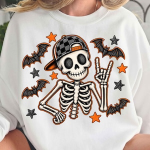 May include: A white crewneck sweatshirt featuring a graphic of a skeleton wearing a black and grey checkered baseball cap. The skeleton is making a hand gesture with fingers extended. The design includes orange and black stars and flying bats.