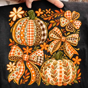 Könnte beinhalten: Eine schwarze Stoffoberfläche mit einem Grafikdruck von zwei Kürbissen und zwei Schleifen. Die Kürbisse und Schleifen sind mit orangefarbenen, weißen und braunen Mustern verziert, darunter Vichy-Karo, Leopardenmuster, Punkte und Blumenmotive.