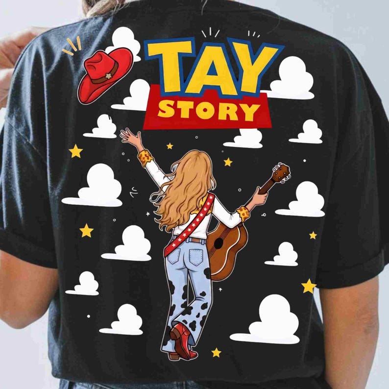 May include: A black t-shirt featuring a graphic print of a person with long blonde hair, wearing a white shirt and cow-print trousers, holding a guitar. The design includes a red cowboy hat, yellow stars, white clouds, and the text TAY STORY.