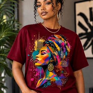 May include: A maroon short-sleeved t-shirt featuring a vibrant, multi-colored graphic print of a person wearing a patterned headwrap and large gold earrings. The artwork incorporates abstract splashes of blue, yellow, pink, and orange paint.