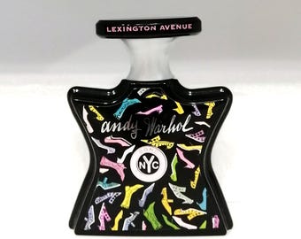 May include: Black perfume bottle with a unique shape, featuring colorful shoe illustrations. The bottle has a black cap with the text "LEXINGTON AVENUE" and the words "andy Warhol" and "NYC" are also visible.