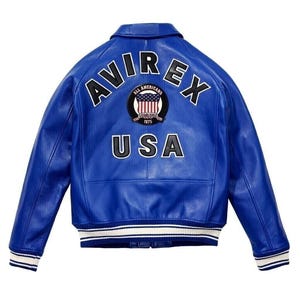 K&ouml;nnte beinhalten: Eine leuchtend blaue Lederjacke mit dem Schriftzug AVIREX in gro&szlig;en schwarzen Buchstaben auf dem oberen R&uuml;cken. Unter dem Text befindet sich ein kreisf&ouml;rmiges Abzeichen mit einem amerikanischen Flaggendesign und den Worten ALL AMERICANS ESTABLISHED 1975. Das Wort USA ist darunter in Schwarz gedruckt. Die Jacke hat einen gerippten Kragen, B&uuml;ndchen und Saum mit wei&szlig;en und blauen Streifen.