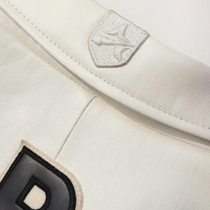 K&ouml;nnte beinhalten: Eine wei&szlig;e Lederjacke mit einem auff&auml;lligen schwarzen Buchstabenaufn&auml;her und einem kleinen, gestickten schildf&ouml;rmigen Emblem am Kragen. Das Emblem zeigt ein stilisiertes grafisches Design, das mit wei&szlig;em Garn auf das wei&szlig;e Leder gestickt ist.