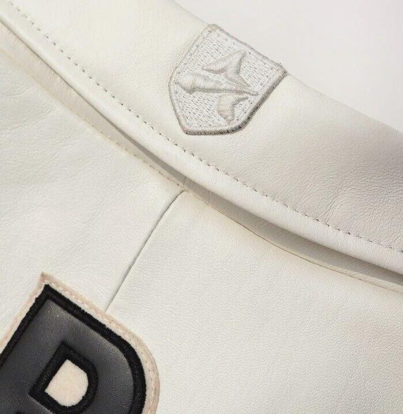 K&ouml;nnte beinhalten: Eine wei&szlig;e Lederjacke mit einem auff&auml;lligen schwarzen Buchstabenaufn&auml;her und einem kleinen, gestickten schildf&ouml;rmigen Emblem am Kragen. Das Emblem zeigt ein stilisiertes grafisches Design, das mit wei&szlig;em Garn auf das wei&szlig;e Leder gestickt ist.