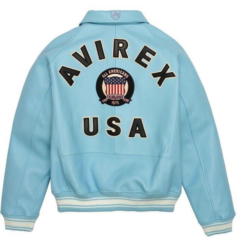 K&ouml;nnte beinhalten: Eine hellblaue Collegejacke mit dem Schriftzug AVIREX und USA in schwarzen Buchstaben auf der R&uuml;ckseite. Ein kreisf&ouml;rmiger Aufn&auml;her in der Mitte zeigt den Text ALL AMERICANS ESTABLISHED 1975 mit einer Grafik der US-Flagge. Die Jacke hat Rippb&uuml;ndchen.