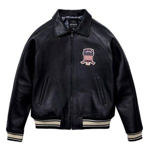 K&ouml;nnte beinhalten: Eine schwarze Lederjacke mit Frontrei&szlig;verschluss, spitzem Kragen und zwei Seitentaschen. Auf der Brust befindet sich ein gestickter Aufn&auml;her mit dem Text AVIREX und einem Schild-Motiv mit der amerikanischen Flagge. Die B&uuml;ndchen und der Saum sind gerippt.