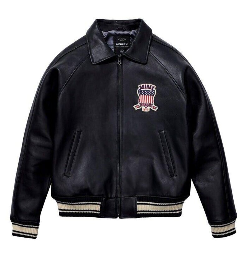 K&ouml;nnte beinhalten: Eine schwarze Lederjacke mit Frontrei&szlig;verschluss, spitzem Kragen und zwei Seitentaschen. Auf der Brust befindet sich ein gestickter Aufn&auml;her mit dem Text AVIREX und einem Schild-Motiv mit der amerikanischen Flagge. Die B&uuml;ndchen und der Saum sind gerippt.