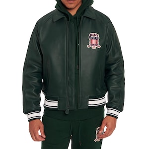 K&ouml;nnte beinhalten: Eine dunkelgr&uuml;ne Leder-Bomberjacke mit klassischem Kragen, Rei&szlig;verschluss vorne und gerippten B&uuml;ndchen mit wei&szlig;en Streifen. Ein Aufn&auml;her auf der linken Brust zeigt den Schriftzug AVIREX mit der amerikanischen Flagge und dem Text ESTABLISHED 1975.