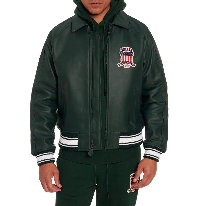 K&ouml;nnte beinhalten: Eine dunkelgr&uuml;ne Leder-Bomberjacke mit klassischem Kragen, Rei&szlig;verschluss vorne und gerippten B&uuml;ndchen mit wei&szlig;en Streifen. Ein Aufn&auml;her auf der linken Brust zeigt den Schriftzug AVIREX mit der amerikanischen Flagge und dem Text ESTABLISHED 1975.