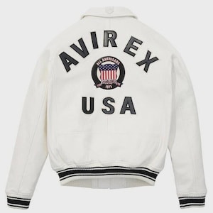 K&ouml;nnte beinhalten: Eine wei&szlig;e Leder-Bomberjacke mit schwarzer Aufschrift AVIREX und USA auf der R&uuml;ckseite. Ein kreisf&ouml;rmiger Aufn&auml;her in der Mitte zeigt den Text ALL AMERICANS ESTABLISHED 1975 mit einem Wappengrafik. Die B&uuml;ndchen und der Saum haben schwarz-wei&szlig;e Streifen.
