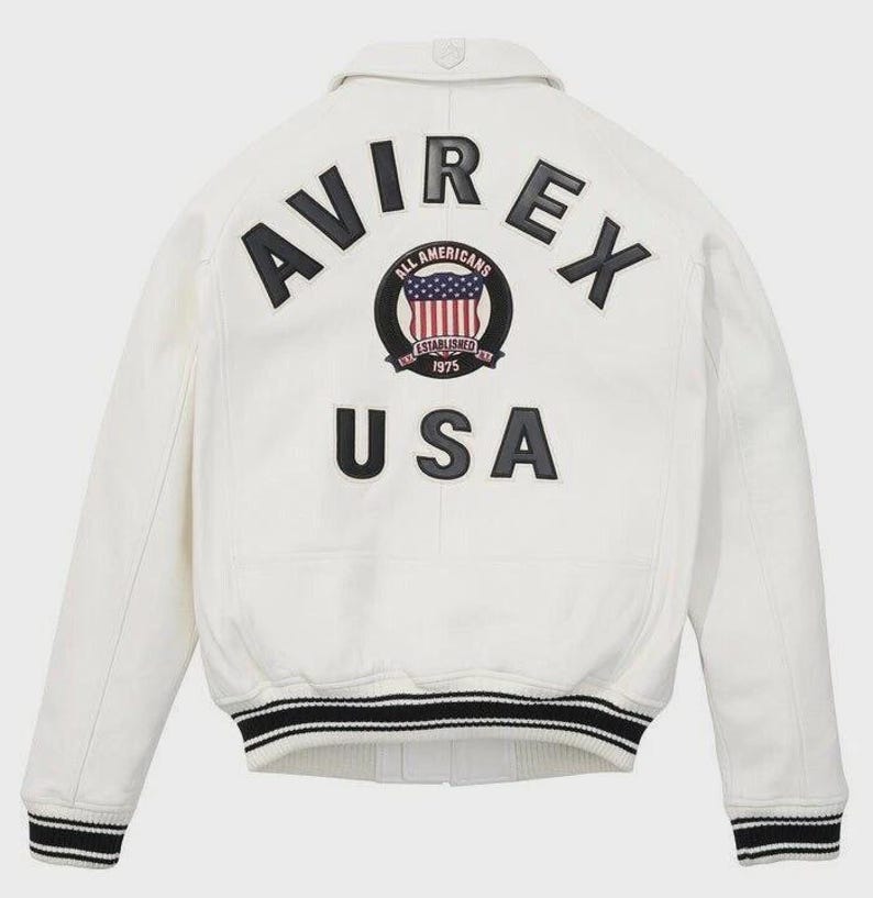 K&ouml;nnte beinhalten: Eine wei&szlig;e Leder-Bomberjacke mit schwarzer Aufschrift AVIREX und USA auf der R&uuml;ckseite. Ein kreisf&ouml;rmiger Aufn&auml;her in der Mitte zeigt den Text ALL AMERICANS ESTABLISHED 1975 mit einem Wappengrafik. Die B&uuml;ndchen und der Saum haben schwarz-wei&szlig;e Streifen.