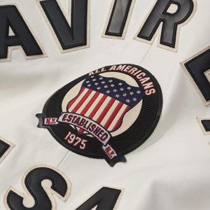 K&ouml;nnte beinhalten: Eine wei&szlig;e Jacke mit gro&szlig;en schwarzen Blockbuchstaben und einem runden gestickten Aufn&auml;her. Der Aufn&auml;her zeigt einen Schild mit der amerikanischen Flagge, den Text ALL AMERICANS, ESTABLISHED 1975 und N.Y. Das Design enth&auml;lt schwarze und wei&szlig;e N&auml;hte.