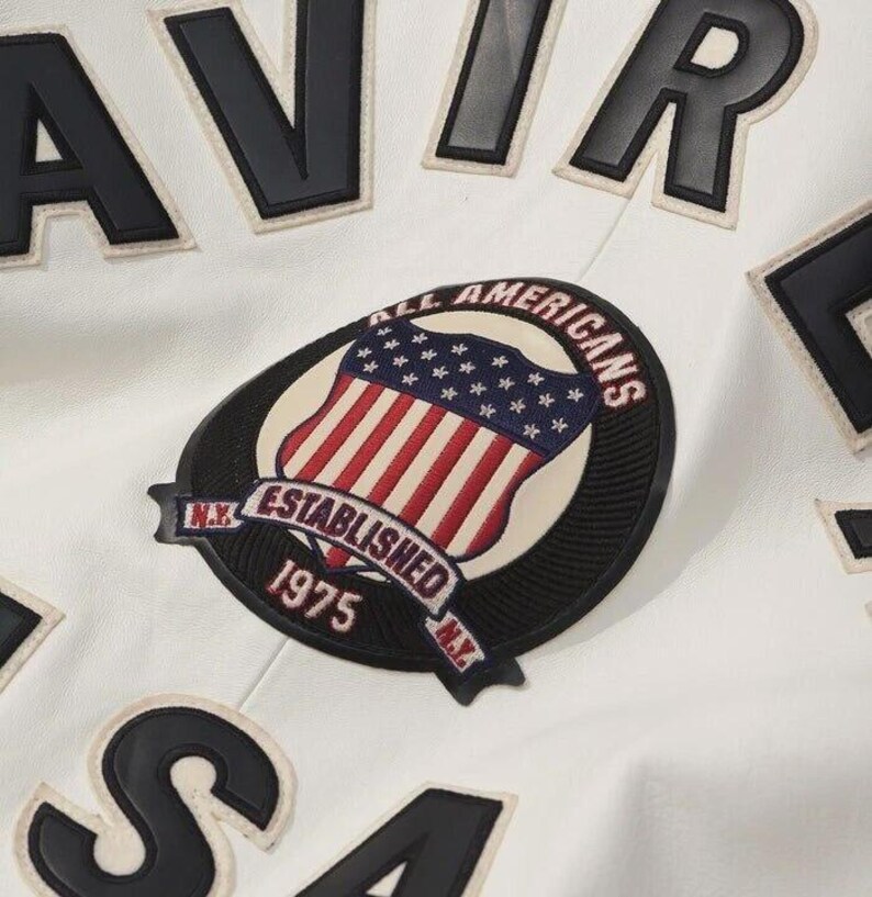 K&ouml;nnte beinhalten: Eine wei&szlig;e Jacke mit gro&szlig;en schwarzen Blockbuchstaben und einem runden gestickten Aufn&auml;her. Der Aufn&auml;her zeigt einen Schild mit der amerikanischen Flagge, den Text ALL AMERICANS, ESTABLISHED 1975 und N.Y. Das Design enth&auml;lt schwarze und wei&szlig;e N&auml;hte.