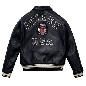 K&ouml;nnte beinhalten: Eine schwarze Leder-Bomberjacke mit dem Schriftzug AVIREX in wei&szlig;en, gebogenen Buchstaben auf dem oberen R&uuml;cken. Darunter befindet sich ein kreisf&ouml;rmiger Aufn&auml;her mit einem amerikanischen Flaggendesign und dem Text ALL AMERICANS ESTABLISHED 1975. Das Wort USA ist in gro&szlig;en wei&szlig;en Buchstaben unten aufgedruckt, erg&auml;nzt durch gestreifte Rippb&uuml;ndchen an &Auml;rmeln und Saum.