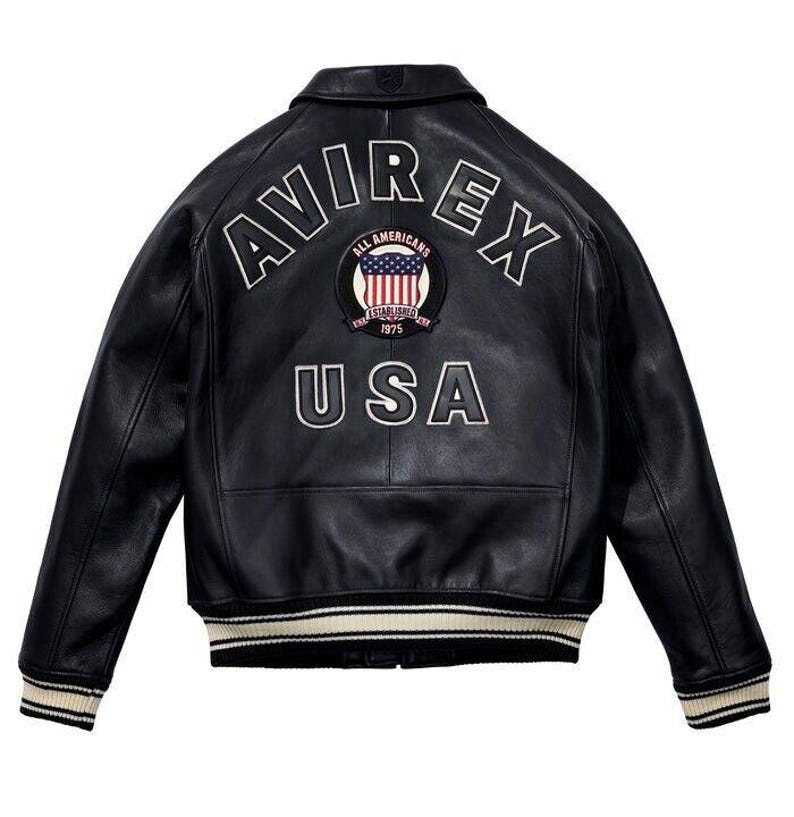 K&ouml;nnte beinhalten: Eine schwarze Leder-Bomberjacke mit dem Schriftzug AVIREX in wei&szlig;en, gebogenen Buchstaben auf dem oberen R&uuml;cken. Darunter befindet sich ein kreisf&ouml;rmiger Aufn&auml;her mit einem amerikanischen Flaggendesign und dem Text ALL AMERICANS ESTABLISHED 1975. Das Wort USA ist in gro&szlig;en wei&szlig;en Buchstaben unten aufgedruckt, erg&auml;nzt durch gestreifte Rippb&uuml;ndchen an &Auml;rmeln und Saum.