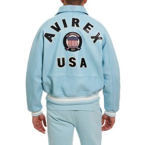 K&ouml;nnte beinhalten: Eine hellblaue Jacke mit geripptem Kragen und B&uuml;ndchen mit wei&szlig;en Streifen. Auf der R&uuml;ckseite befindet sich der schwarze Schriftzug AVIREX und USA sowie ein kreisf&ouml;rmiger Aufn&auml;her mit einer US-Flagge und dem Text ALL AMERICANS ESTABLISHED 1975.