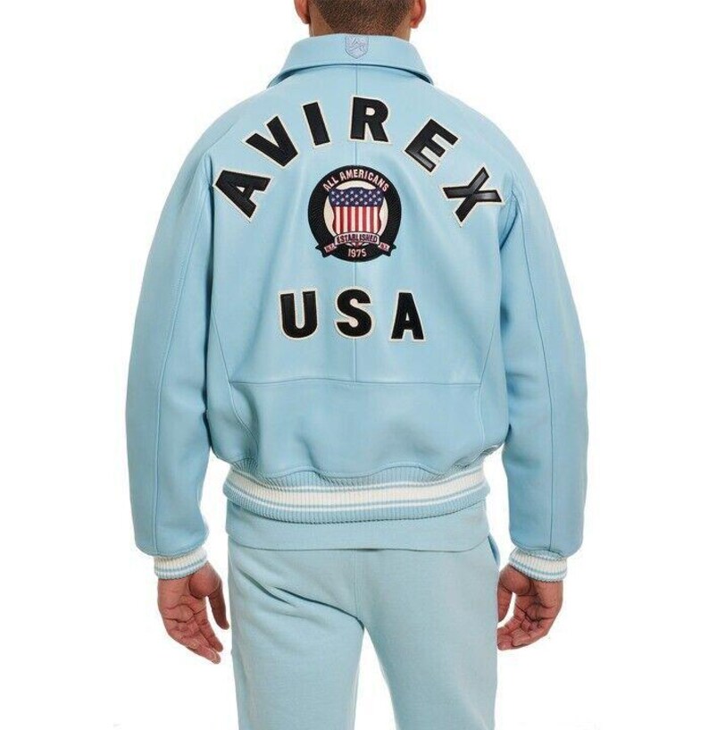 K&ouml;nnte beinhalten: Eine hellblaue Jacke mit geripptem Kragen und B&uuml;ndchen mit wei&szlig;en Streifen. Auf der R&uuml;ckseite befindet sich der schwarze Schriftzug AVIREX und USA sowie ein kreisf&ouml;rmiger Aufn&auml;her mit einer US-Flagge und dem Text ALL AMERICANS ESTABLISHED 1975.