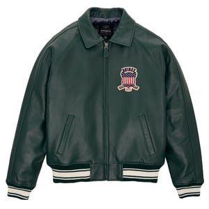 K&ouml;nnte beinhalten: Eine dunkelgr&uuml;ne Lederjacke mit durchgehendem Frontrei&szlig;verschluss, klassischem Kragen sowie gerippten B&uuml;ndchen und Saum mit wei&szlig;en Streifen. Ein gestickter Aufn&auml;her auf der linken Brust zeigt den Text AVIREX &uuml;ber einem Wappen mit US-Flagge.