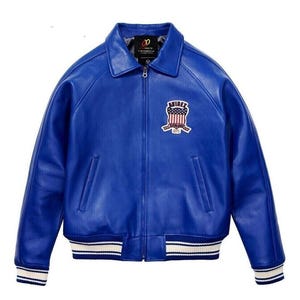 K&ouml;nnte beinhalten: Eine leuchtend blaue Lederjacke mit Frontrei&szlig;verschluss, klassischem Kragen und zwei Seitentaschen. Auf der Brust befindet sich ein gestickter Aufn&auml;her mit dem Text AVIREX und einem Schild-Motiv. Die &Auml;rmelb&uuml;ndchen und der Saum sind wei&szlig;-blau gestreift.