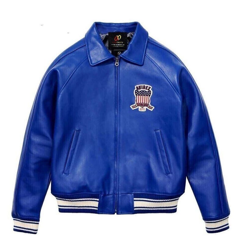 K&ouml;nnte beinhalten: Eine leuchtend blaue Lederjacke mit Frontrei&szlig;verschluss, klassischem Kragen und zwei Seitentaschen. Auf der Brust befindet sich ein gestickter Aufn&auml;her mit dem Text AVIREX und einem Schild-Motiv. Die &Auml;rmelb&uuml;ndchen und der Saum sind wei&szlig;-blau gestreift.