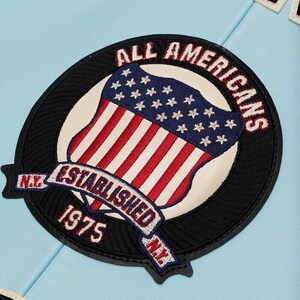 K&ouml;nnte beinhalten: Ein kreisf&ouml;rmiger gestickter Aufn&auml;her auf hellblauem Untergrund. Der Aufn&auml;her zeigt einen Schild mit roten und wei&szlig;en Streifen sowie einem blauen Feld mit wei&szlig;en Sternen. Der Text lautet ALL AMERICANS, ESTABLISHED 1975 und N.Y. in Stickerei.