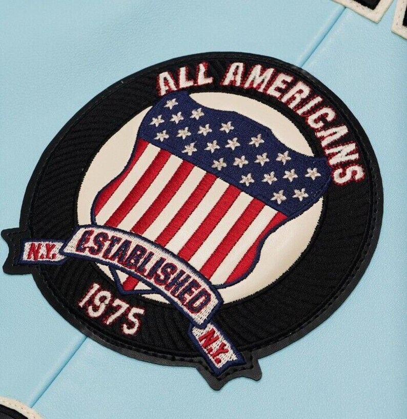 K&ouml;nnte beinhalten: Ein kreisf&ouml;rmiger gestickter Aufn&auml;her auf hellblauem Untergrund. Der Aufn&auml;her zeigt einen Schild mit roten und wei&szlig;en Streifen sowie einem blauen Feld mit wei&szlig;en Sternen. Der Text lautet ALL AMERICANS, ESTABLISHED 1975 und N.Y. in Stickerei.