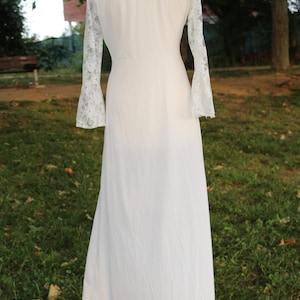 May include: A floor-length white gown featuring long, sheer lace sleeves with flared cuffs. The garment has a solid white bodice and a flowing skirt, displayed on a mannequin. The lace sleeves exhibit a detailed floral pattern throughout the fabric.