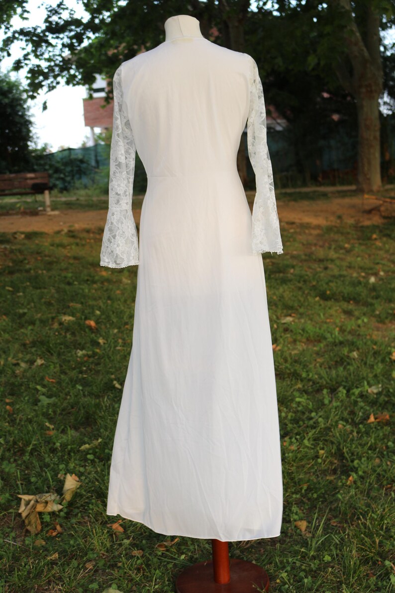 May include: A floor-length white gown featuring long, sheer lace sleeves with flared cuffs. The garment has a solid white bodice and a flowing skirt, displayed on a mannequin. The lace sleeves exhibit a detailed floral pattern throughout the fabric.