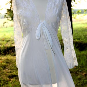 May include: A white long-sleeved robe featuring a sheer lace bodice with a deep V-neckline and a front tie closure. The garment has long, flared lace sleeves and a flowing, semi-sheer skirt that extends to the floor, displayed on a mannequin.
