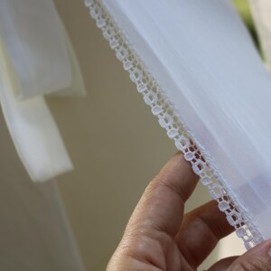 May include: A sheer white fabric garment featuring a delicate, intricate white lace trim along the edge. The material is lightweight and semi-transparent, showcasing a fine weave and a clean, bright white color throughout the textile piece.