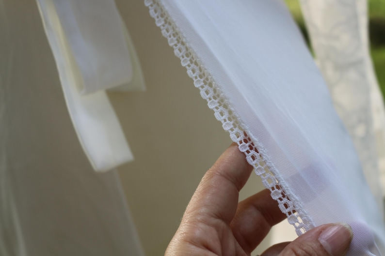 May include: A sheer white fabric garment featuring a delicate, intricate white lace trim along the edge. The material is lightweight and semi-transparent, showcasing a fine weave and a clean, bright white color throughout the textile piece.