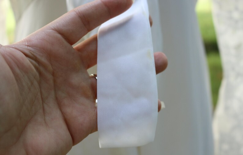 May include: A smooth, white fabric ribbon or strap with a subtle sheen. The material is opaque and features a clean, straight edge, appearing as a component of a larger garment or accessory, captured in a clear, focused view of the textile texture.