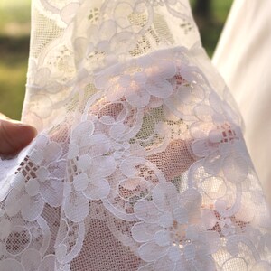 May include: A white lace fabric featuring a detailed floral pattern with intricate scalloped edges and a sheer, lightweight mesh construction. The delicate textile displays a repeating botanical motif with interconnected petals and openwork.