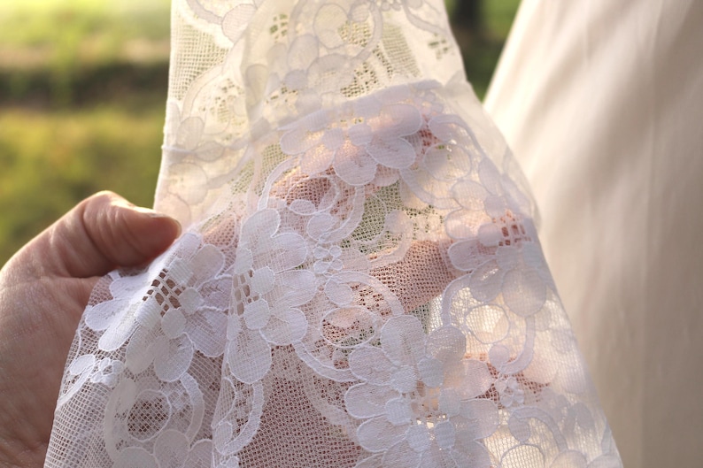May include: A white lace fabric featuring a detailed floral pattern with intricate scalloped edges and a sheer, lightweight mesh construction. The delicate textile displays a repeating botanical motif with interconnected petals and openwork.